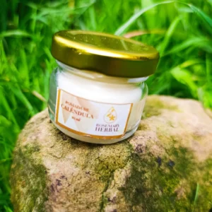 Frasco pequeño de pomada de caléndula sobre piedra, rodeado de pasto verde, marca Rosemary Herbal.